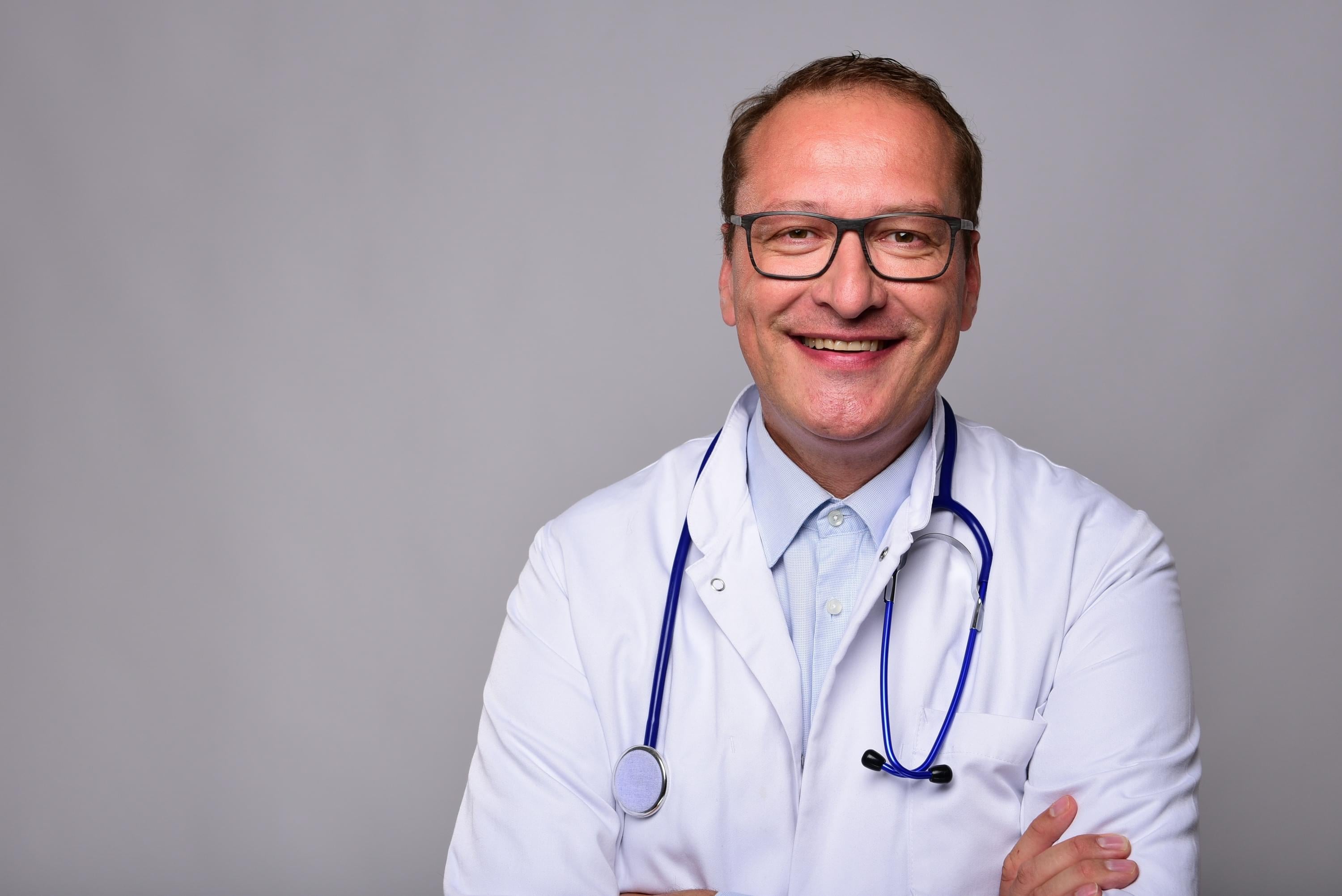 Dr. Johannes Link standing on the right against a grey studio background, stethoscope around his neck
